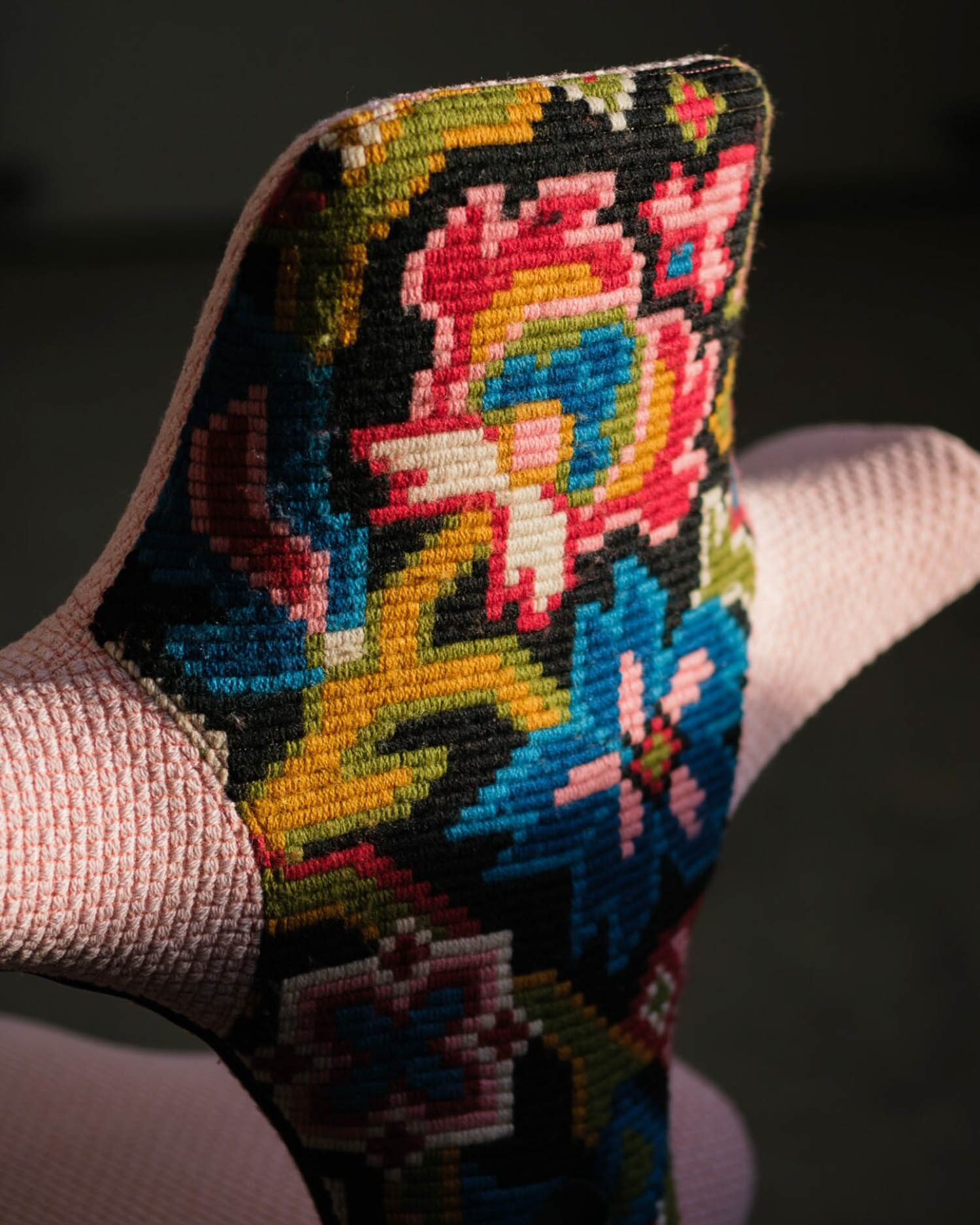 Close-up of a chair with a textured light pink fabric and a backrest featuring a colorful floral needlepoint embroidery pattern.
