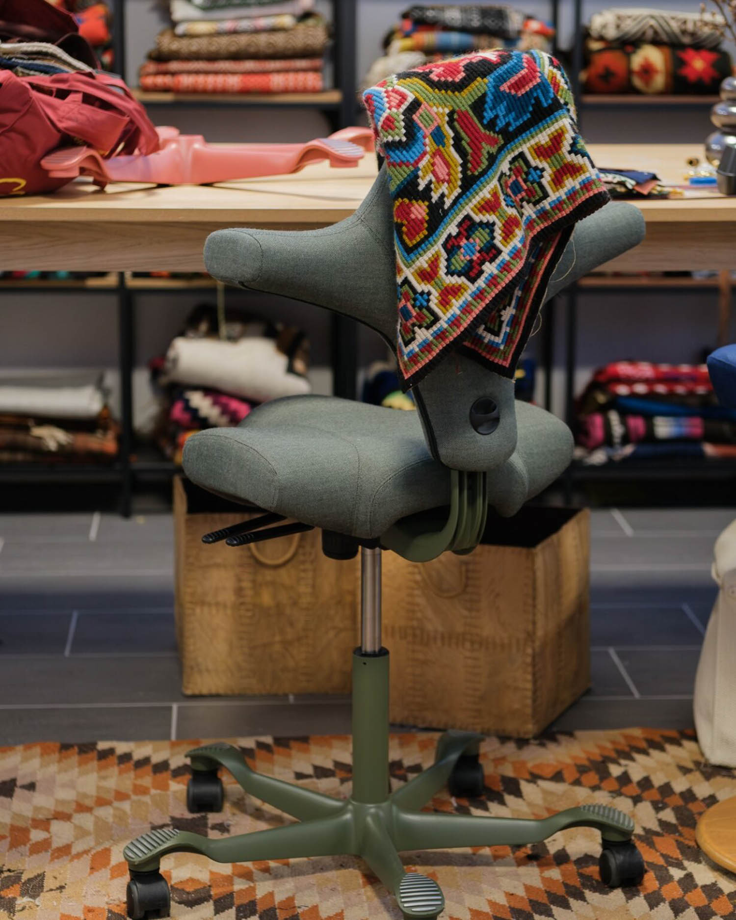 A gray office chair with a colorful patterned textile draped over the backrest, placed in front of shelves filled with folded fabrics.