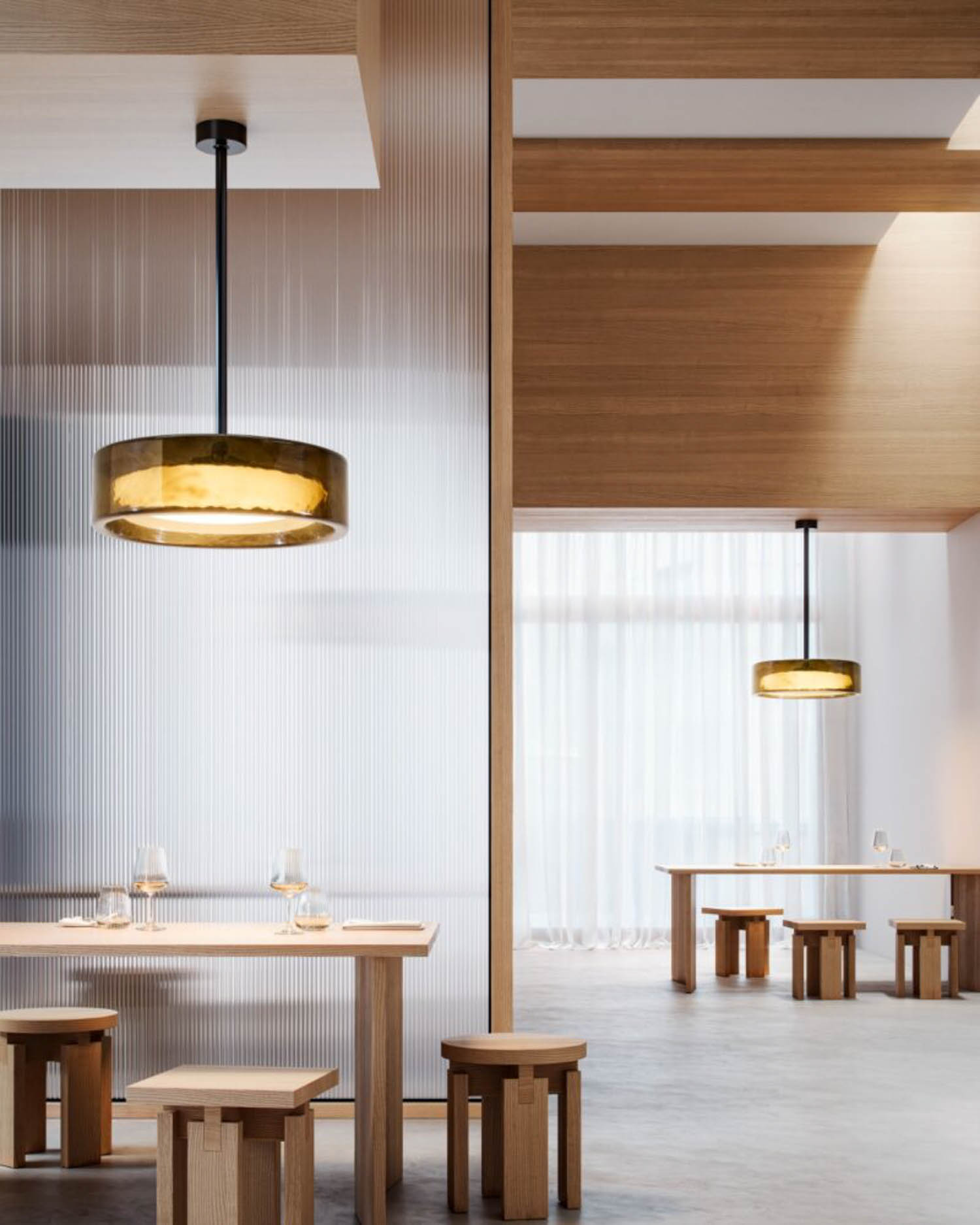 Modern dining area with wooden tables and stools, glassware set on tables, pendant lights, and natural light from tall windows.