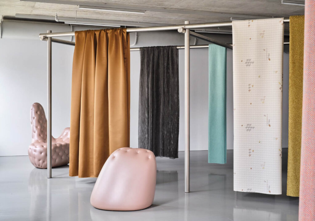 Several colorful fabric panels hang from a metal frame in a modern room with a pink, textured chair and matching ottoman on a shiny floor.