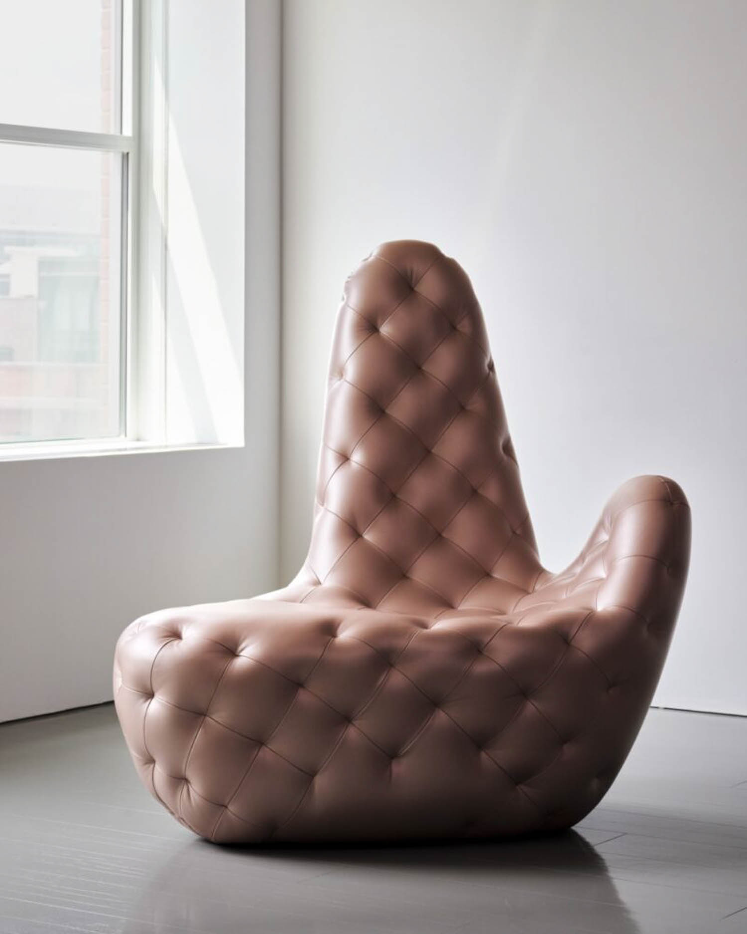 A large, tufted chair upholstered in light brown leather sits in a minimal room next to a window, shaped with a high, rounded backrest and smooth curved lines.
