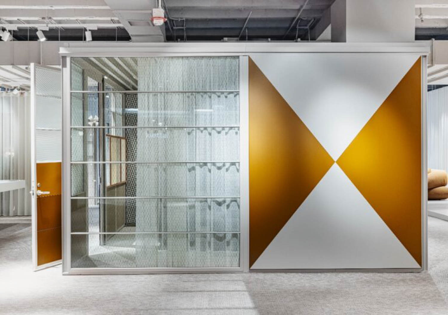 A modern office pod with a glass door and wall on the left and a geometric gold and white design on the right panel, set in an open indoor space.