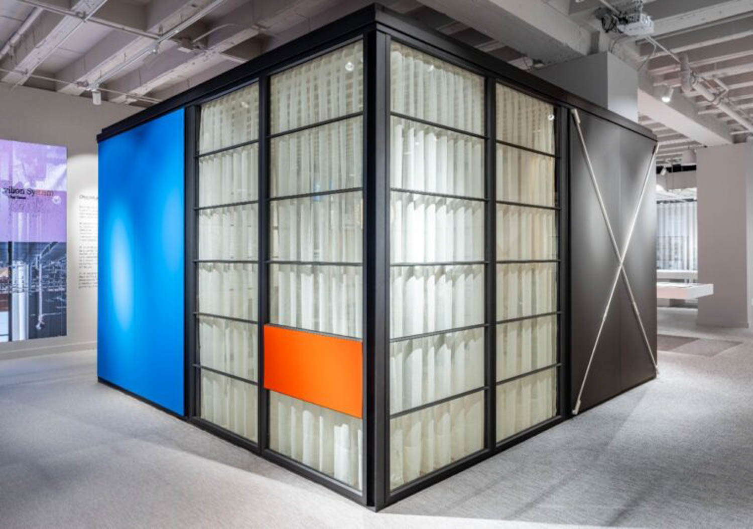 A modern glass-walled room with black metal frames, blue and orange panels, and vertical white cylinders inside, located in a brightly lit exhibition space.