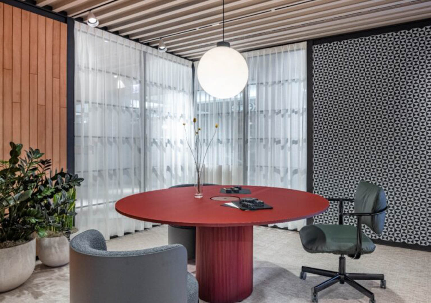 Modern office space with a round red table, two chairs, potted plants, patterned accent wall, sheer curtains, and a spherical ceiling light.