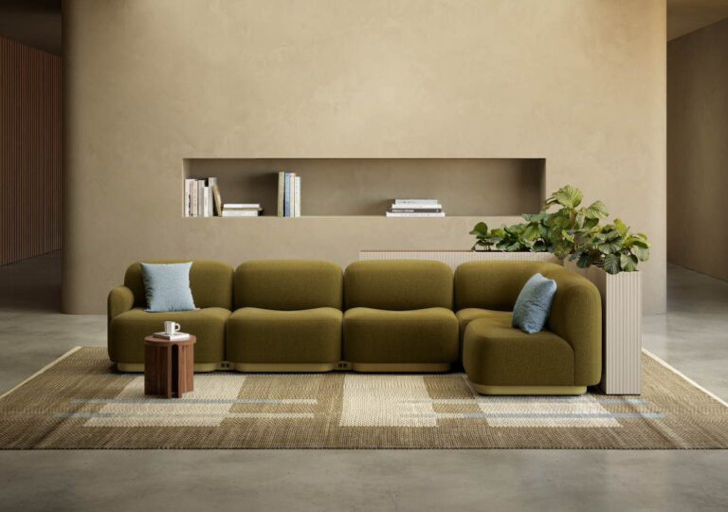 A modern living room with an olive green sectional sofa, two blue cushions, a small round wooden table, a large plant, and a built-in wall shelf with books against a beige wall.