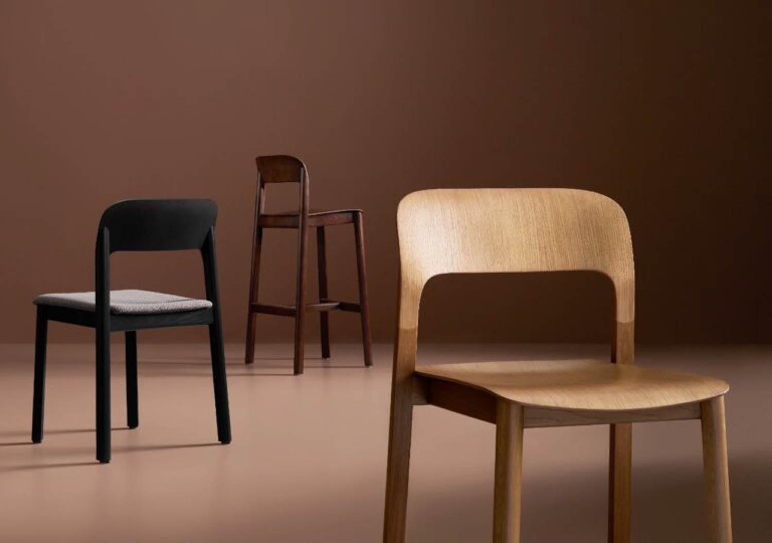 Three modern wooden chairs—one light wood, one black with a gray seat, and one dark brown—are arranged against a brown background.