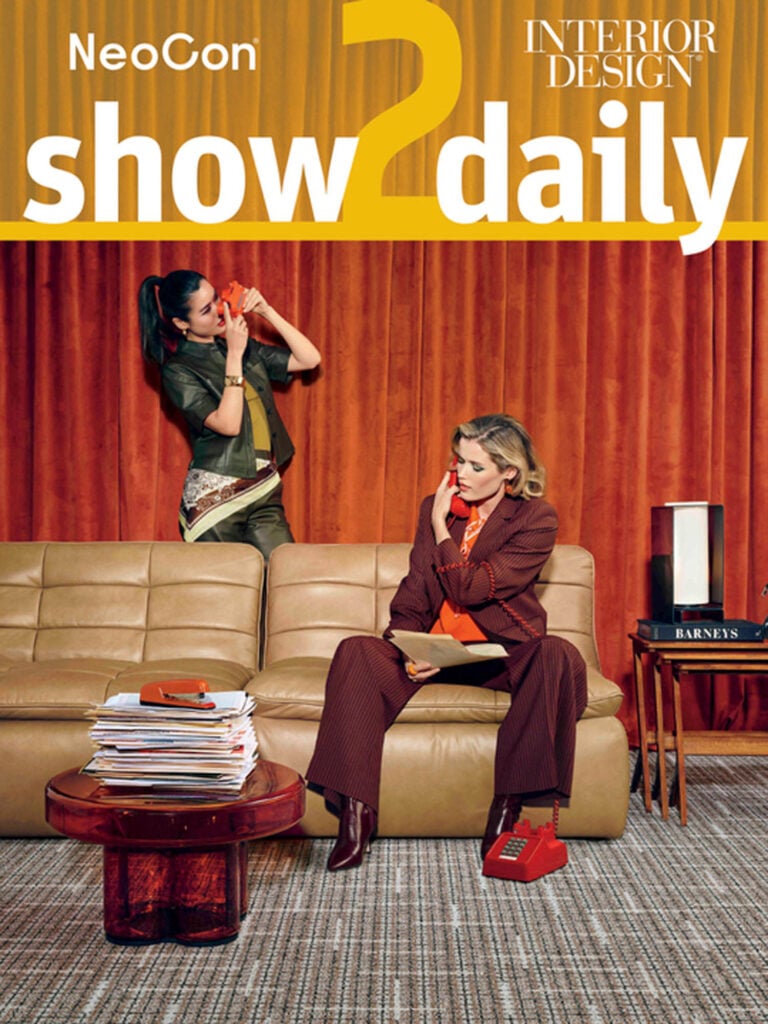 Two women sit and stand near a tan sofa in front of orange curtains; one talks on a red phone, the other sits with a phone, next to a stack of magazines and a box labeled "Barneys.