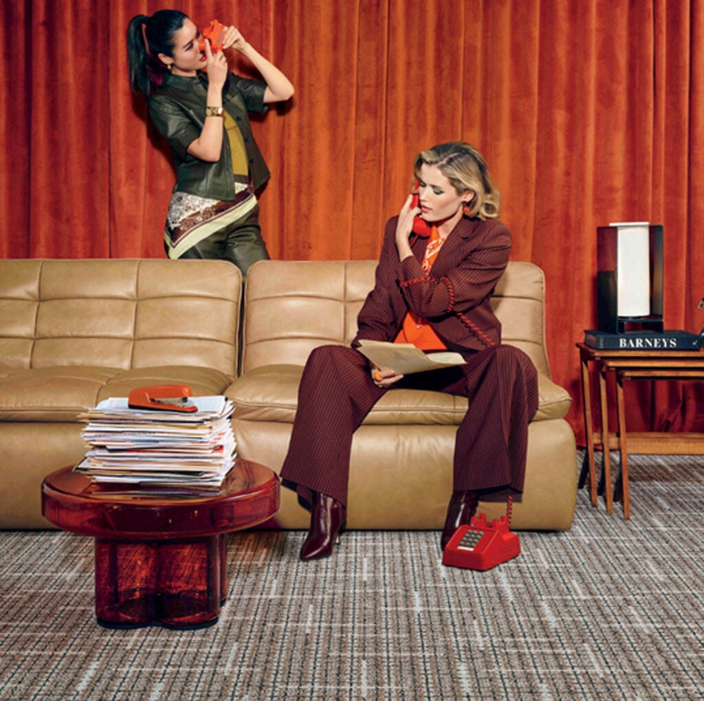 Two women sit and stand near a tan sofa in front of orange curtains; one talks on a red phone, the other sits with a phone, next to a stack of magazines and a box labeled "Barneys.