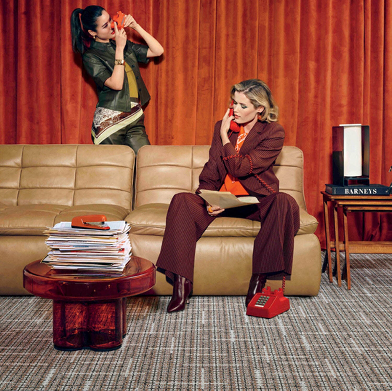 Two women sit and stand near a tan sofa in front of orange curtains; one talks on a red phone, the other sits with a phone, next to a stack of magazines and a box labeled "Barneys.