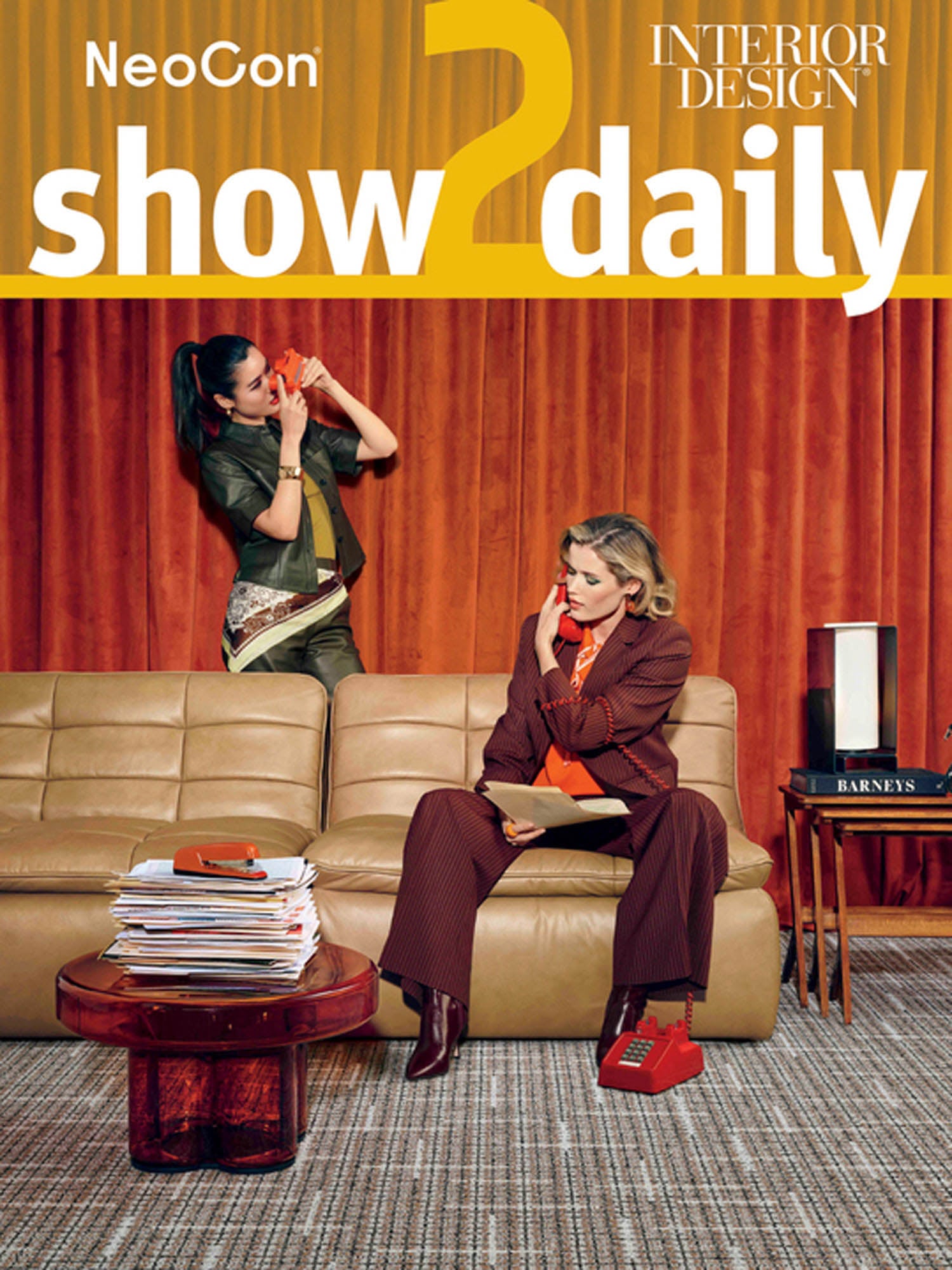 Two women sit and stand near a tan sofa in front of orange curtains; one talks on a red phone, the other sits with a phone, next to a stack of magazines and a box labeled "Barneys.