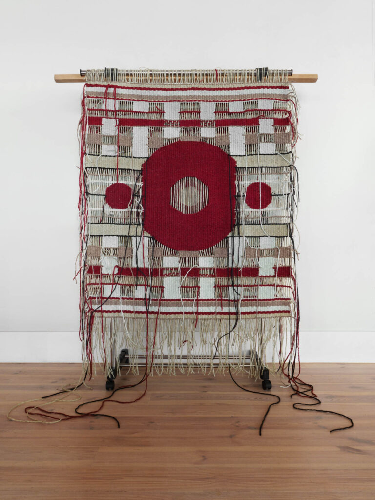 A woven textile art piece with red geometric shapes and loose threads hangs on a wooden rod, displayed against a white wall and wooden floor.