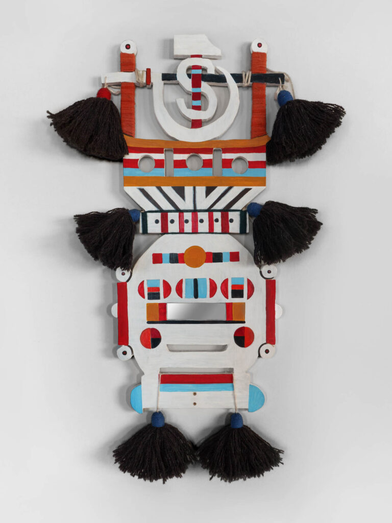 A geometric wooden mask painted in red, blue, black, and white with circular and rectangular cutouts, decorated with dark tassels on the sides and bottom.