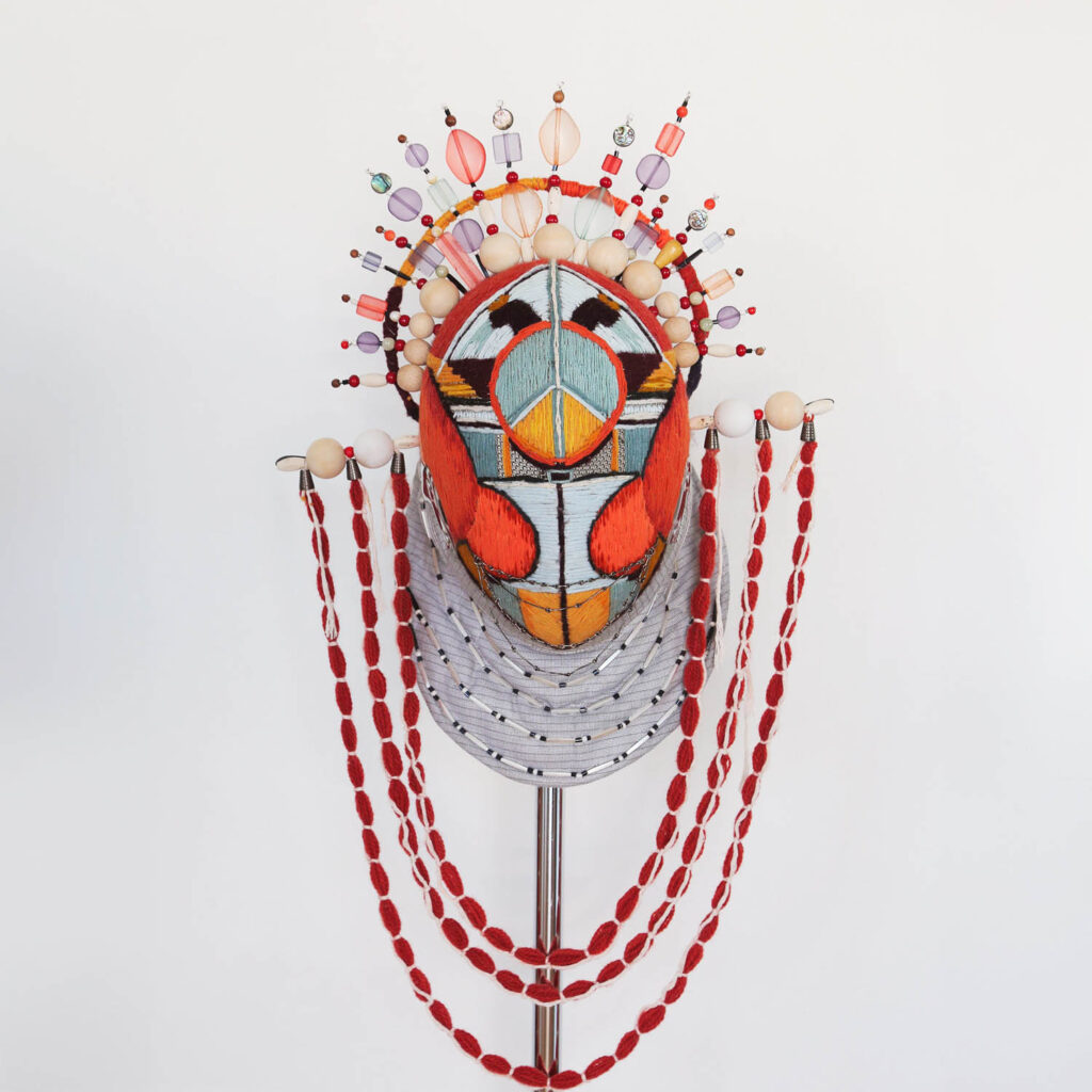 A colorful, ornate mask with geometric patterns, beaded embellishments, and red bead garlands draped on both sides, displayed on a stand against a plain white background.