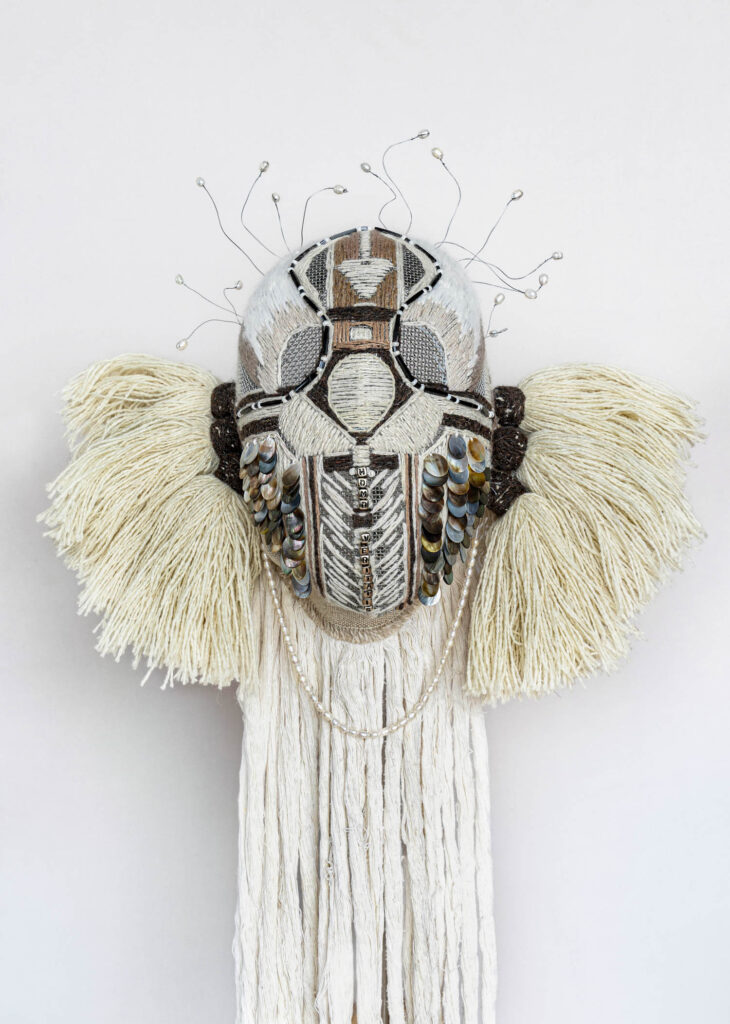 A mixed media mask featuring a beaded geometric design, metallic discs, cream-colored yarn wings, and long hanging strands against a plain background.