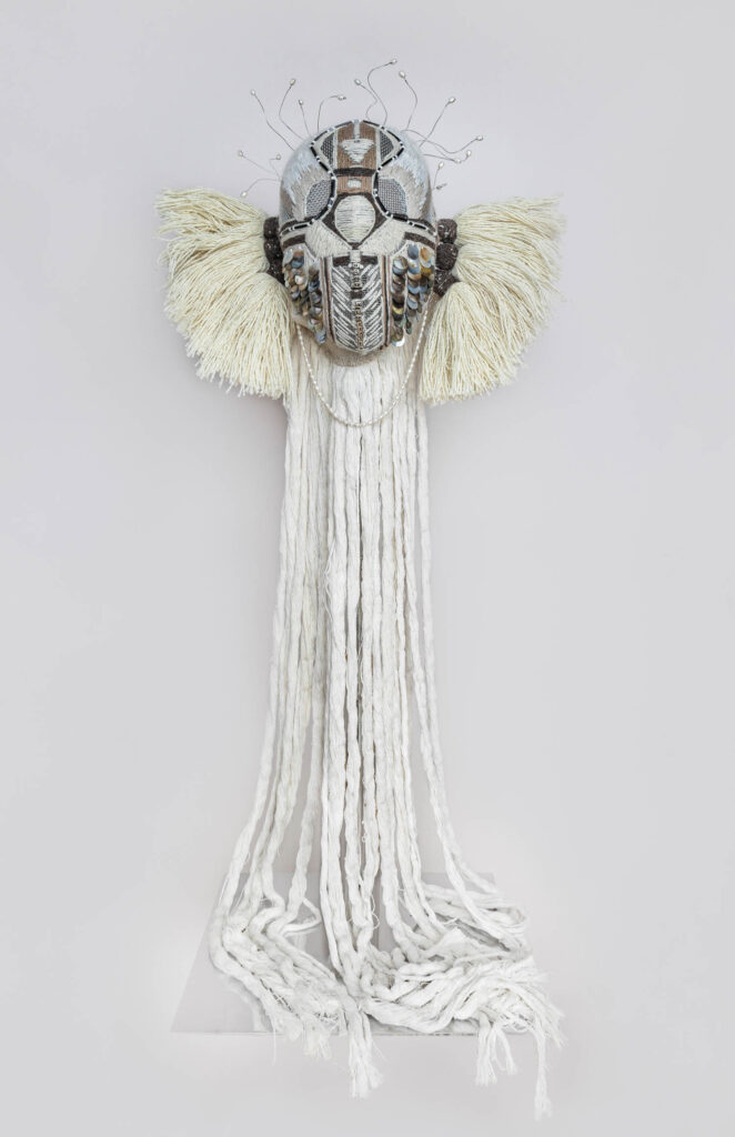 A decorative mask with metallic elements, woven textures, and long white fabric strands hanging down, mounted on a plain light background.