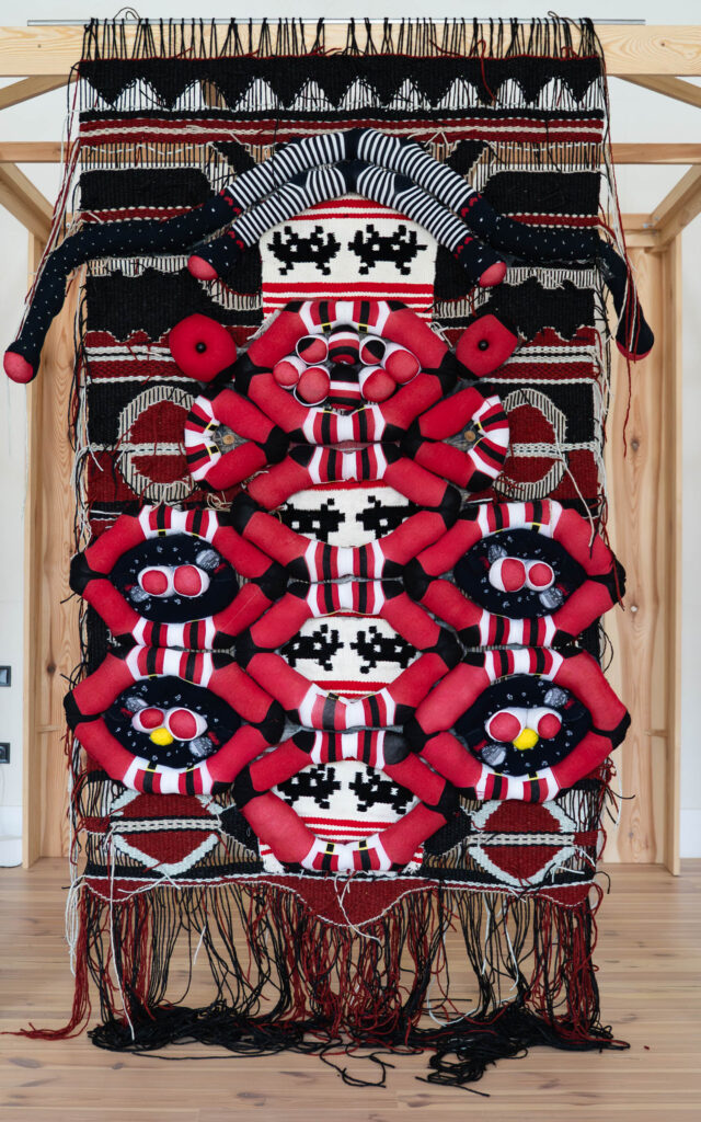 A large woven textile artwork hangs on a wooden frame, featuring geometric red, black, and white patterns with layered soft sculptural elements and loose hanging threads.