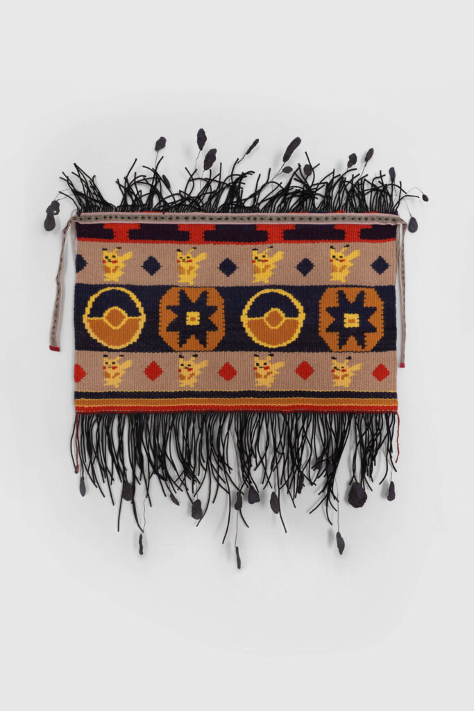 Rectangular woven textile with geometric and circular patterns, red diamonds, black fringe, and black knotted cords hanging from the bottom edge.