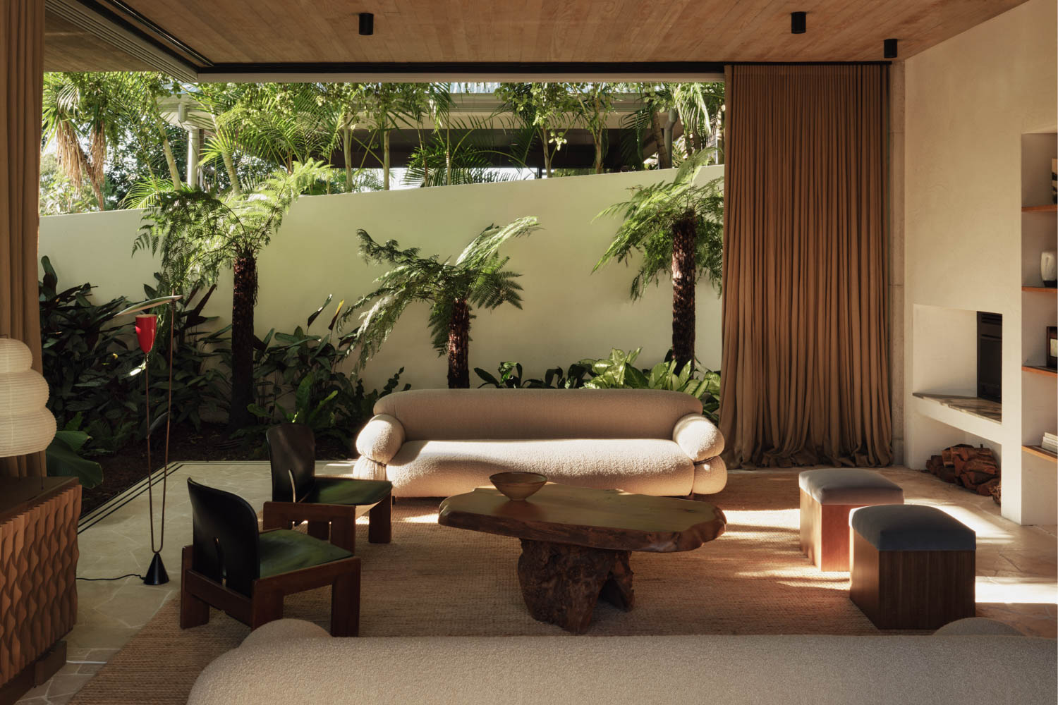 A modern living room with neutral furniture, a wooden coffee table, and large glass doors opening to a lush garden with palm trees and greenery.
