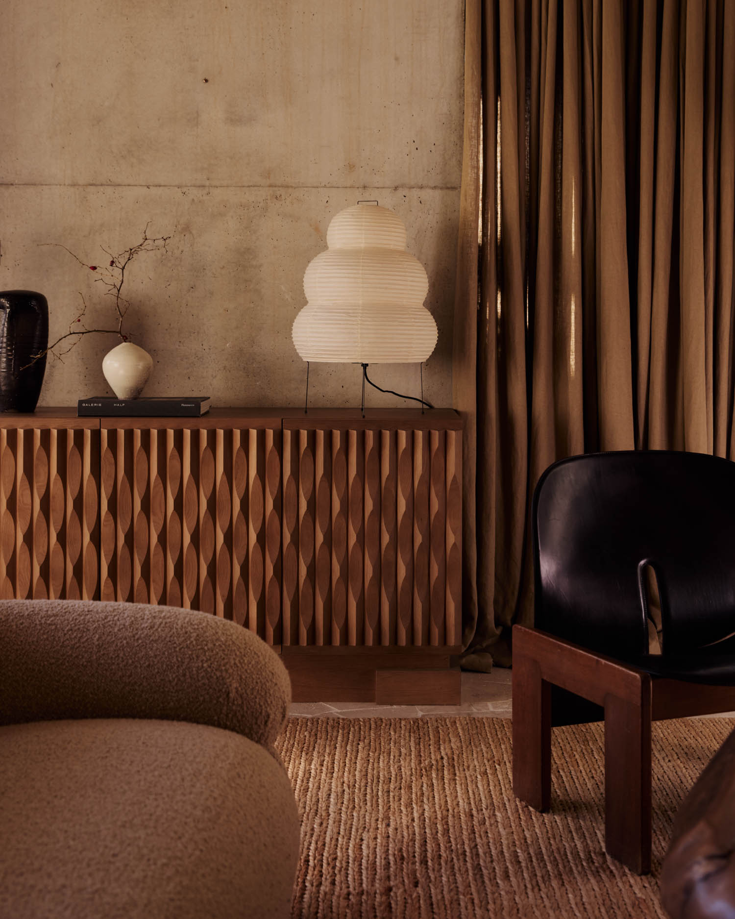 A modern living room with a textured wooden sideboard, a layered paper lamp, a black chair, neutral-toned sofa, rug, and floor-length brown curtains against a concrete wall.