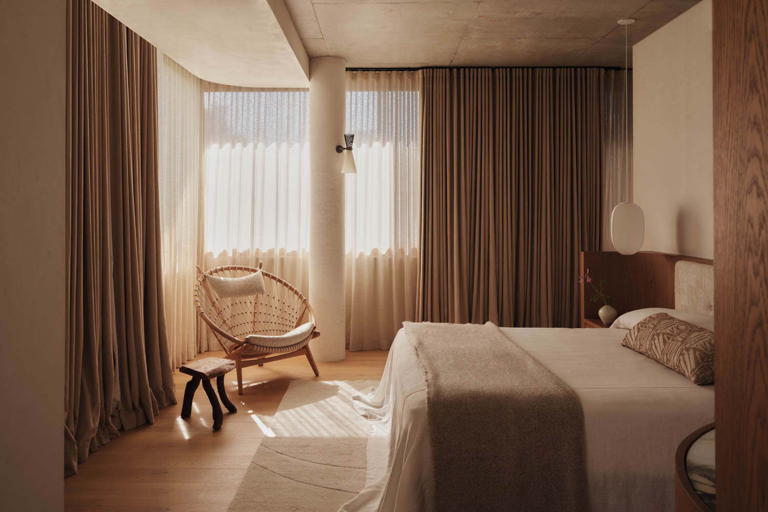 Bedroom with a double bed, neutral bedding, wooden chair, large windows with sheer curtains, and warm natural light.