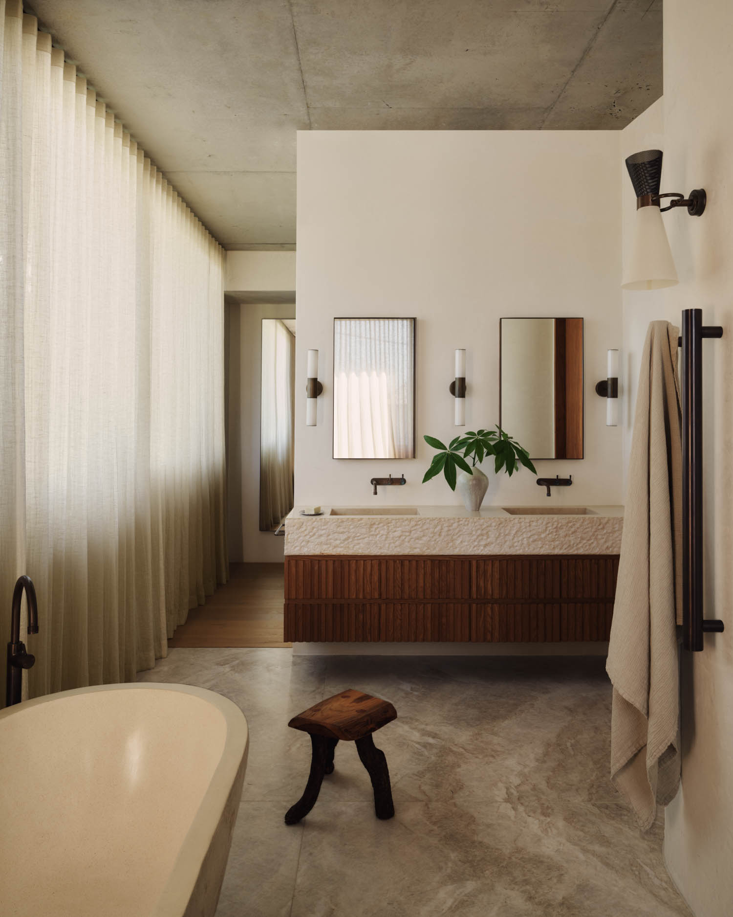 Modern bathroom with a freestanding tub, double sink vanity with two mirrors, wall-mounted faucets, a small wooden stool, and light curtains covering a large window.