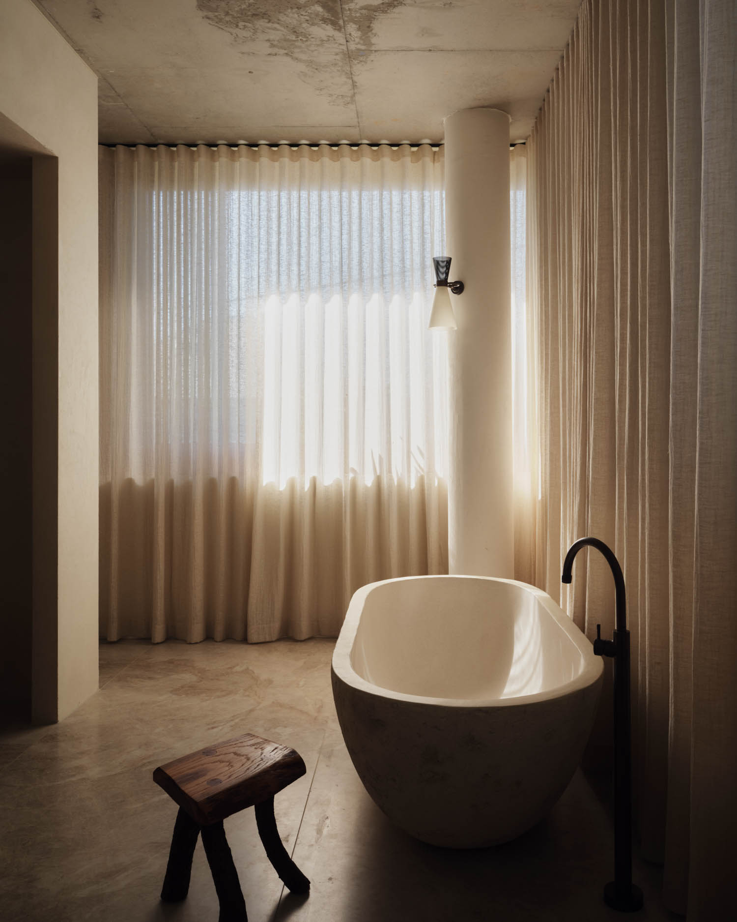 A modern bathroom with a freestanding oval bathtub, a wooden stool, floor-to-ceiling curtains, and a wall-mounted light fixture.