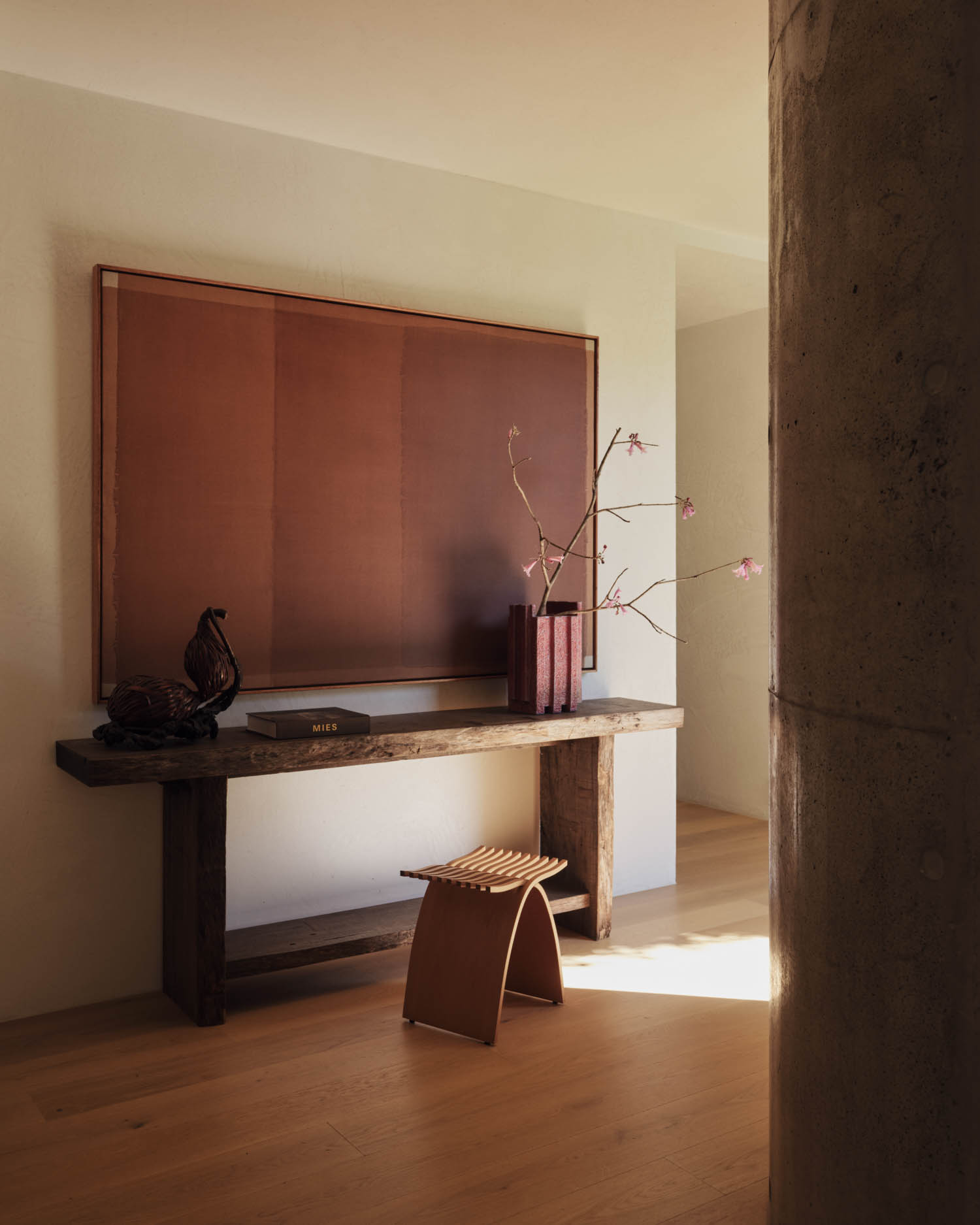 A minimalist interior with a wooden console table, decorative objects, a small stool, a large abstract artwork, and a concrete pillar in the foreground.