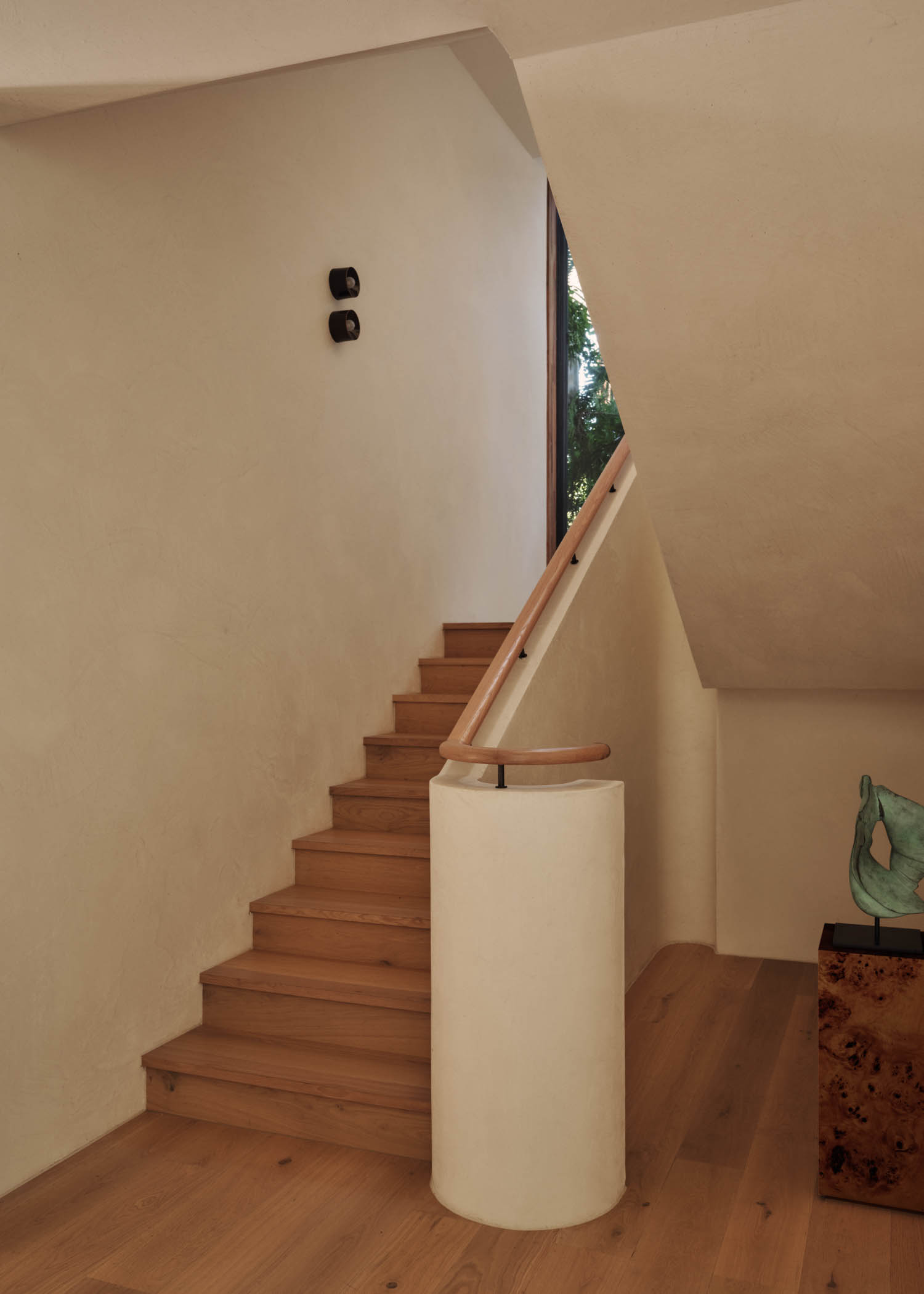 A wooden staircase with a light-colored wall and a simple handrail, leading up beside a window; modern, minimalist interior design.