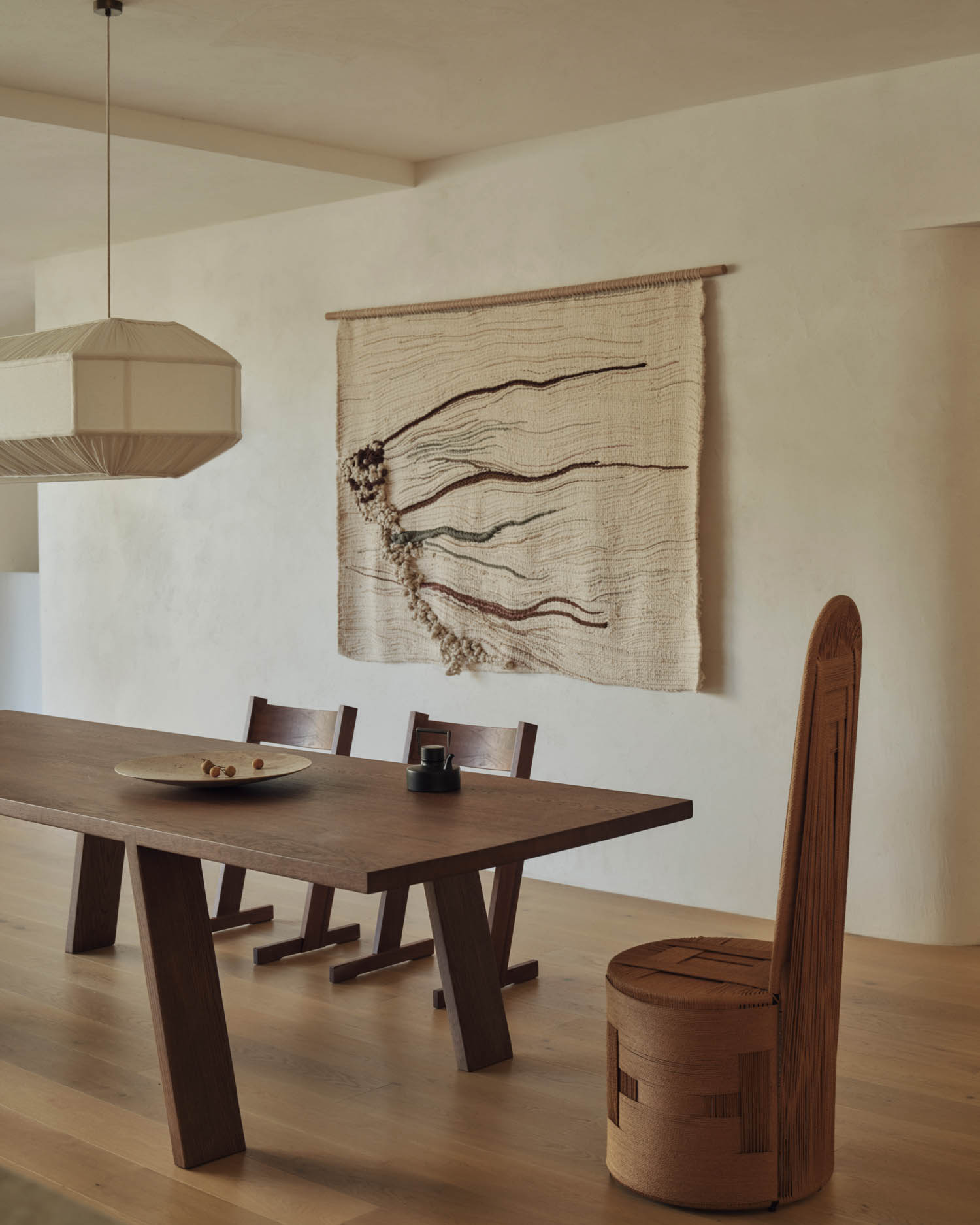 Minimalist dining room with a wooden table, mismatched chairs, a plate and cup, pendant light, and a textured wall tapestry featuring abstract lines.