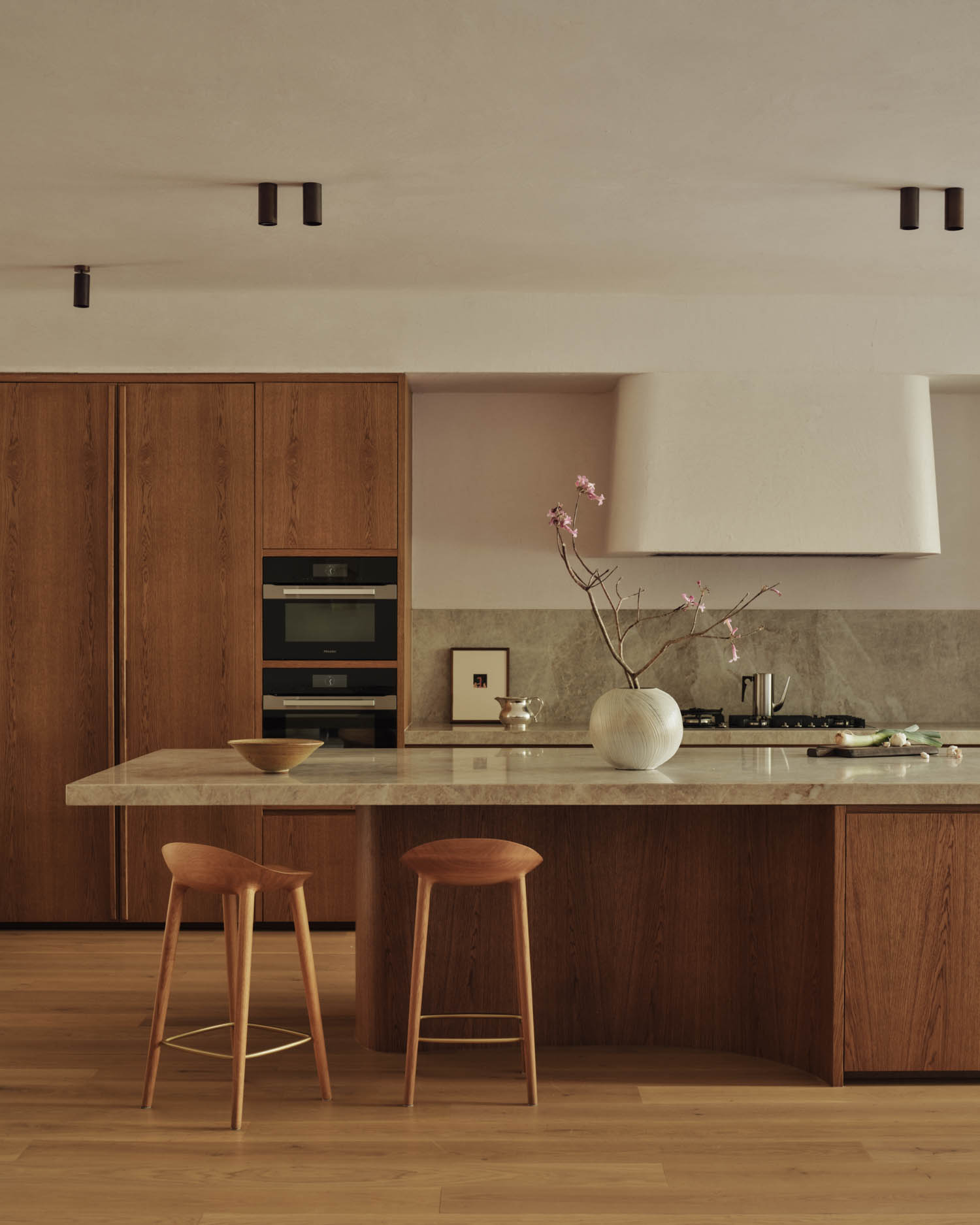 Minimalist kitchen with wood cabinetry, marble island, two wooden stools, built-in ovens, a vase with branches, and neutral-colored walls and flooring.