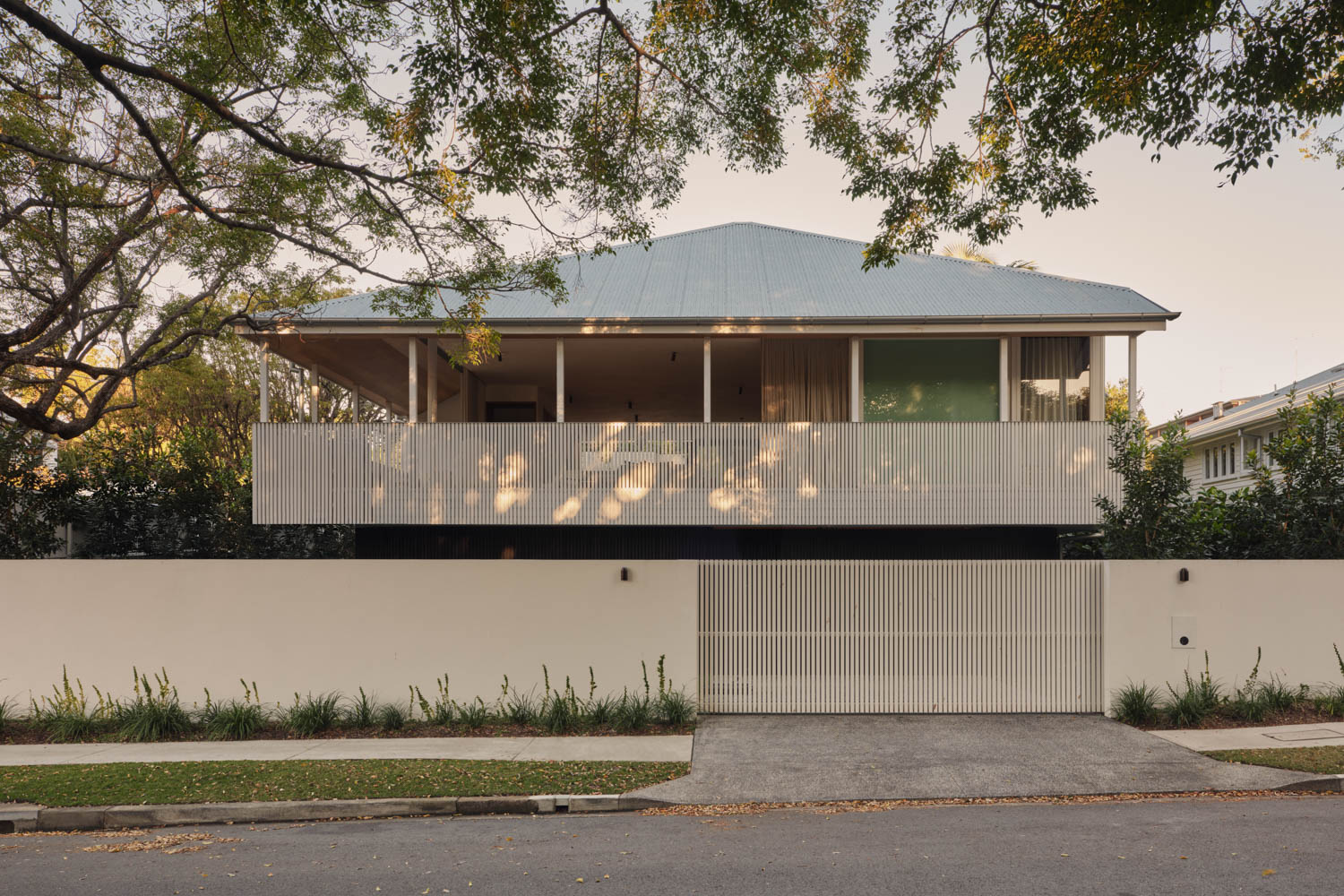 Modern two-story house with a large front balcony, white exterior, metal roof, and a gated driveway, surrounded by trees and a simple garden.
