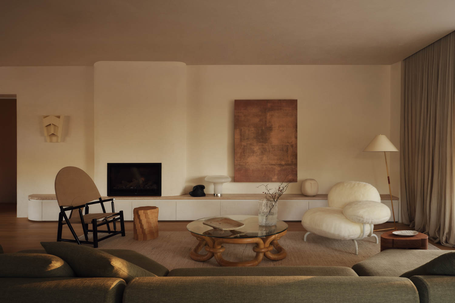 Modern living room with neutral tones, low white cabinetry, abstract wall art, unique sculptural furniture, a fireplace, and sheer curtains, all softly lit by natural light.