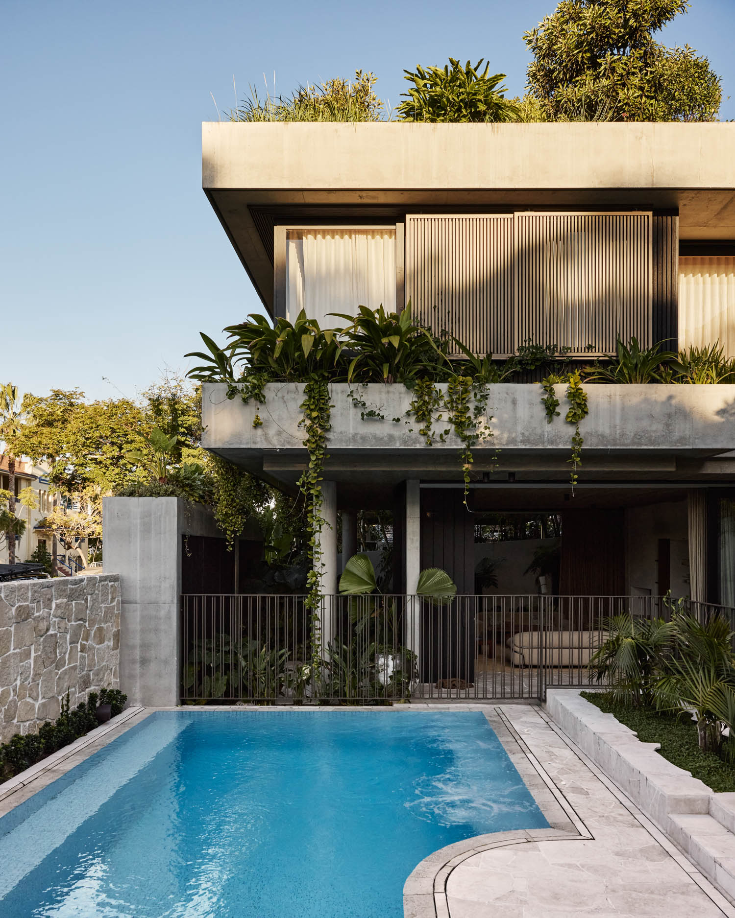 Modern concrete house with large windows and lush greenery on balconies, overlooking a backyard swimming pool with a stone wall and sunlit patio area.