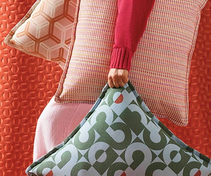 A person wearing a red sweater holds three decorative pillows with geometric patterns against a textured red background. "NeoCon show daily 1" and "Interior Design" text overlays the image.