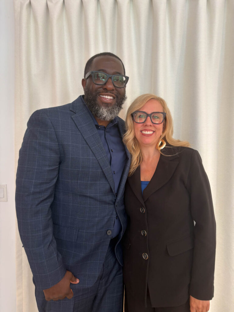 Two people stand side by side in front of white curtains, both wearing glasses and business attire, smiling at the camera.