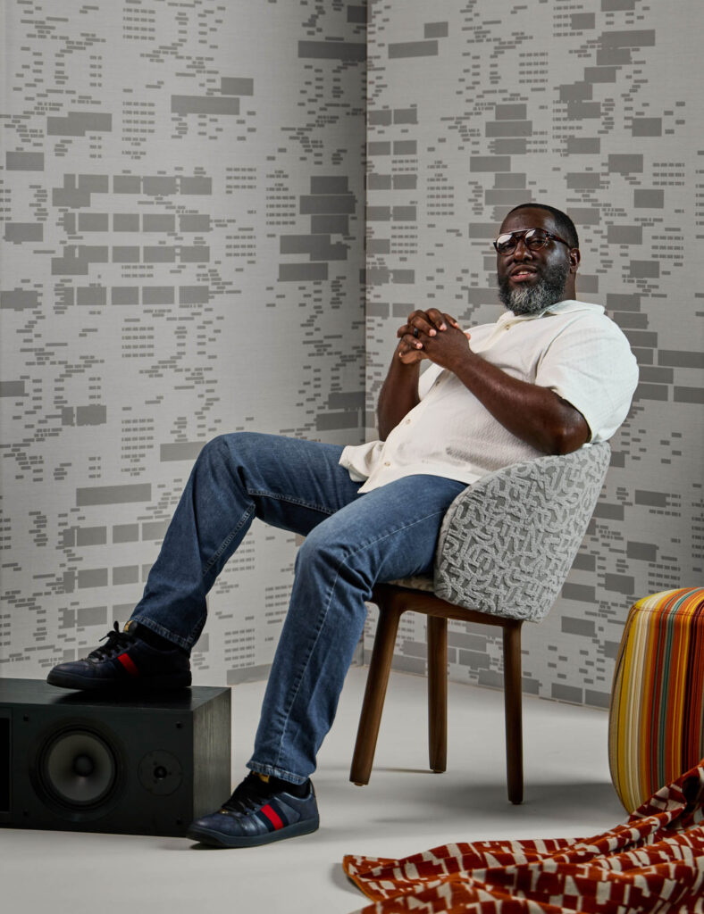 A man wearing glasses, a white shirt, and jeans sits on a patterned chair in a modern room with geometric wallpaper, a speaker, and a striped fabric on the floor.