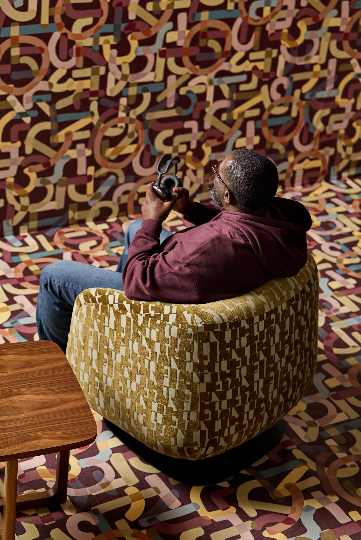A man in a maroon hoodie sits in a patterned swivel chair, holding headphones, surrounded by an abstract, colorful patterned floor and wall.