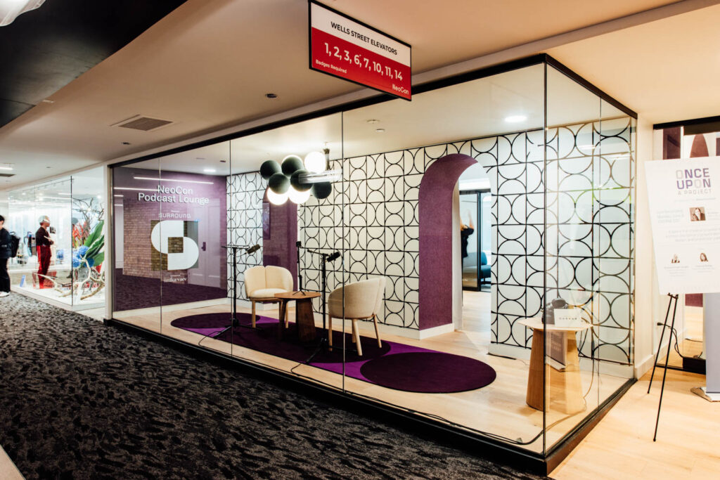 Glass-walled podcast lounge with two chairs, a table, microphones, modern decor, patterned walls, and a ceiling light fixture in a hallway setting.