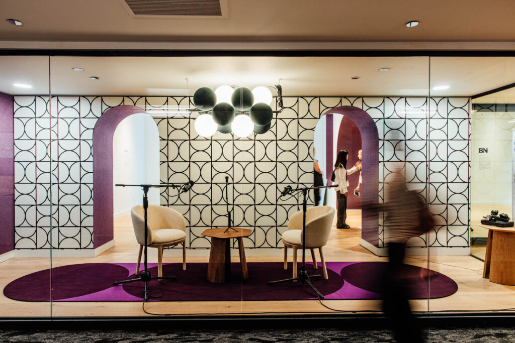 Glass-walled room with two chairs, microphones, and a small table on a purple carpet, set against a geometric-patterned wall with arches. A person walks past outside.