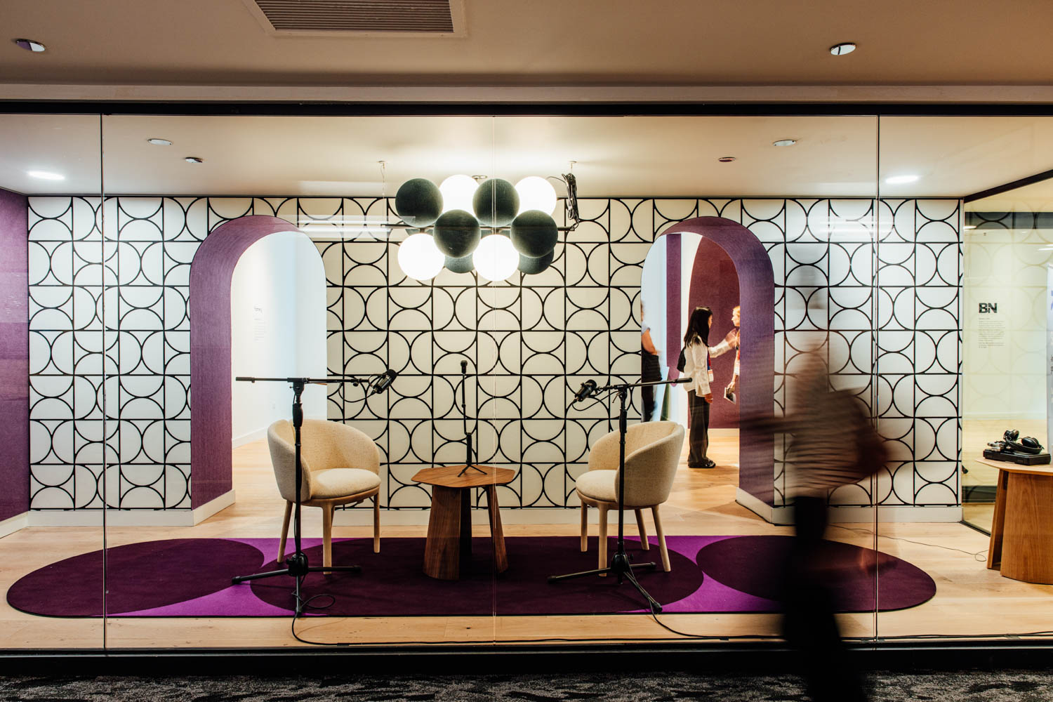 Glass-walled room with two chairs, microphones, and a small table on a purple carpet, set against a geometric-patterned wall with arches. A person walks past outside.