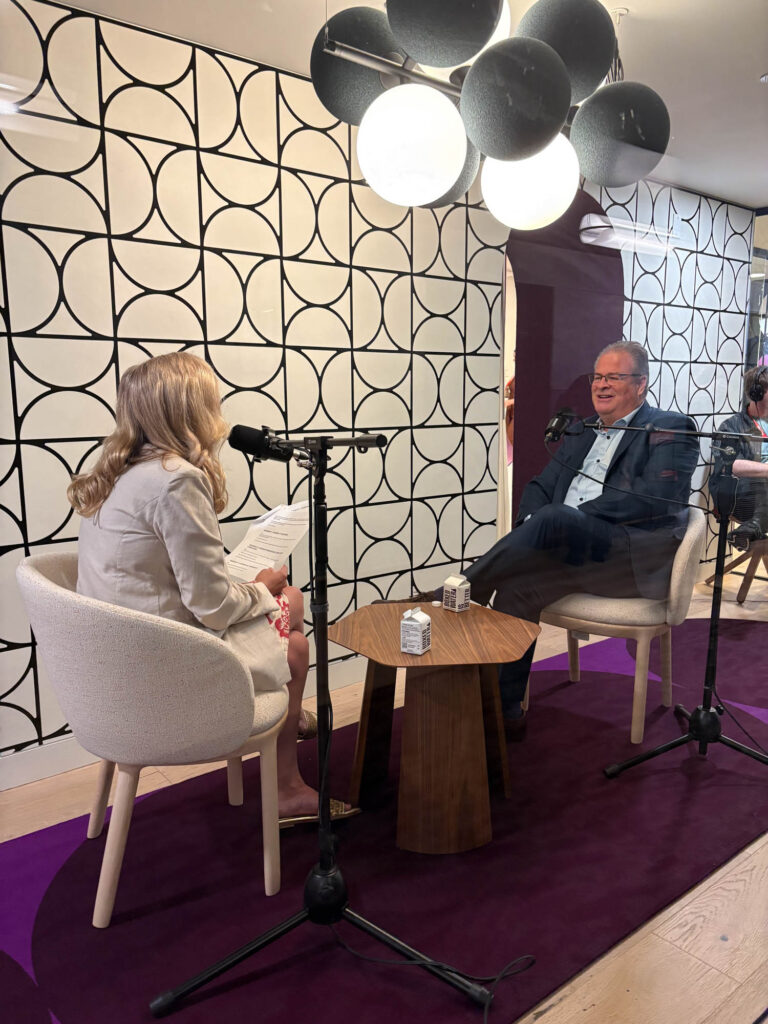 Two people sit across from each other in a modern studio, speaking into microphones, likely recording an interview. A geometric-patterned wall and round overhead lights are visible.