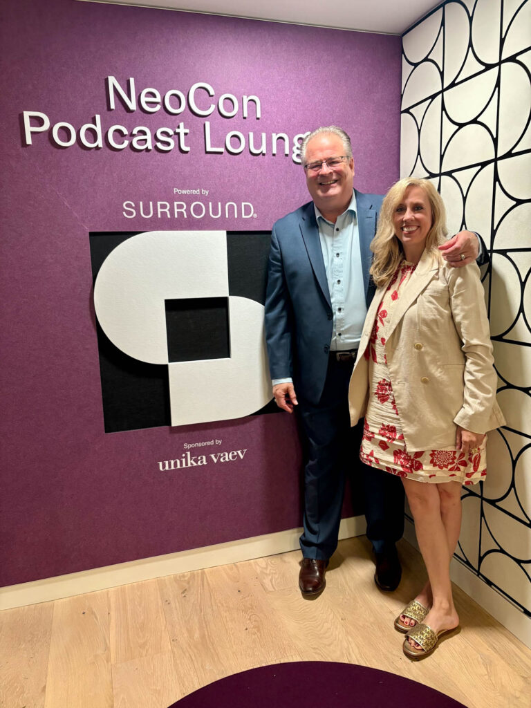 Two people stand and smile in front of a purple wall with the text "NeoCon Podcast Lounge powered by Surround, sponsored by unika vaev.