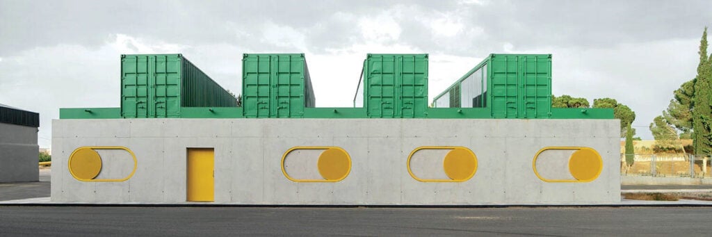 A modern building with a gray concrete base featuring yellow circular windows and door, and three large green shipping containers on the roof.