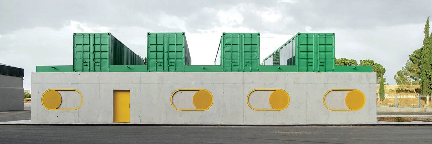 A modern building with a gray concrete base featuring yellow circular windows and door, and three large green shipping containers on the roof.