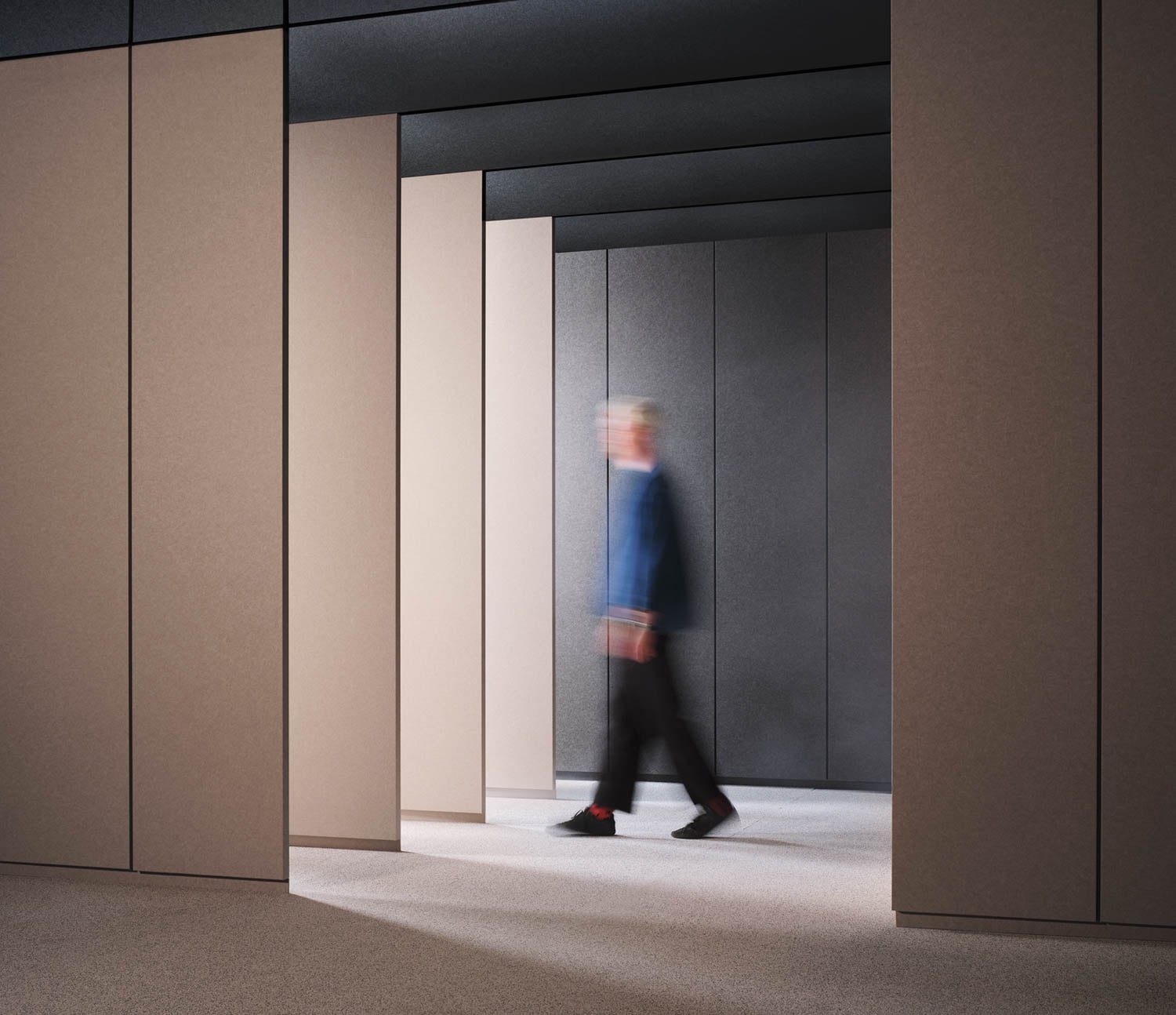 A person in motion walks through a modern, minimalistic hallway with beige and dark gray walls.