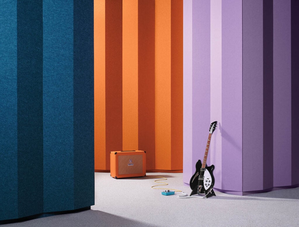 A black electric guitar, effects pedal, and orange amplifier are placed on a light floor in front of tall, multicolored vertical panels in blue, orange, and purple.