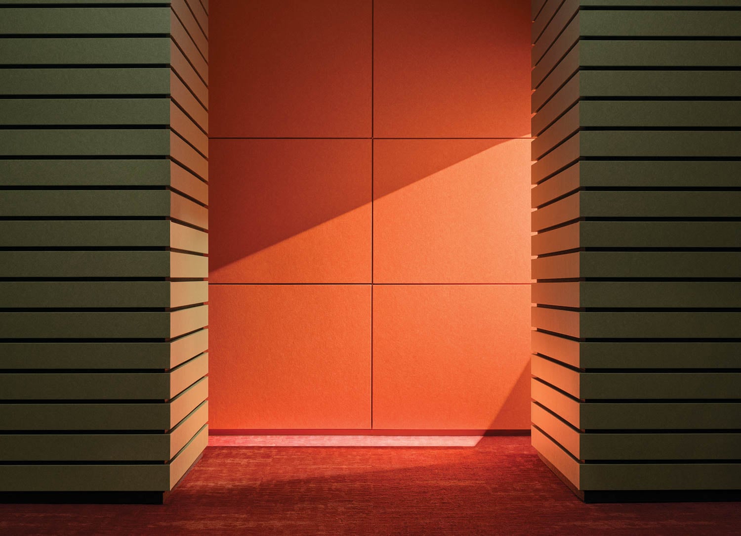 A geometric space with green horizontal-slatted walls and an orange paneled backdrop, illuminated by diagonal sunlight on a red carpeted floor.