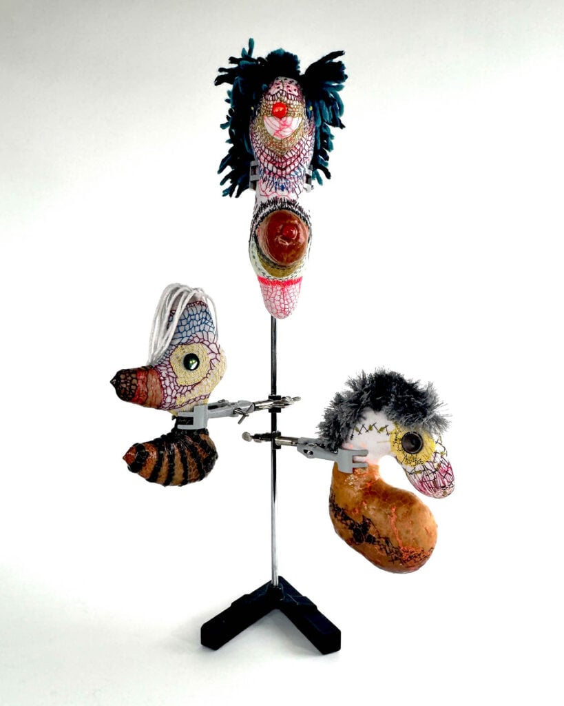 Three colorful, textured, abstract heads with yarn hair by Wolfgang Woerner are mounted on a metal stand against a white background.