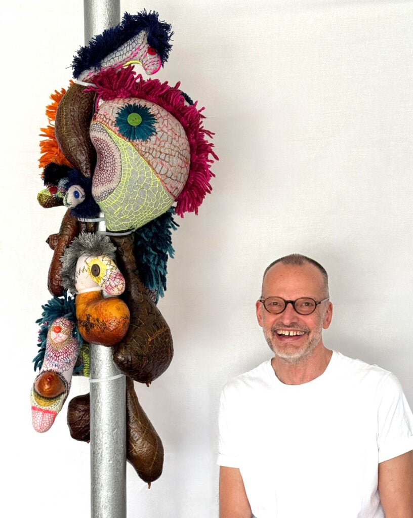 A smiling man in glasses and a white t-shirt sits beside a pole adorned with colorful, textured fabric and object sculptures against a plain white background—an artful scene by Wolfgang Woerner.