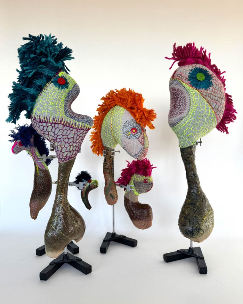 Five colorful, abstract, bird-like sculptures by Wolfgang Woerner with textured surfaces and button eyes are displayed on stands against a plain white background.